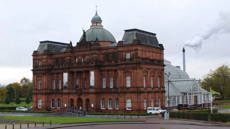 A large, red sandstone Victorian building with a dome and statues stands in front of a modern glasshouse. Smoke rises from a chimney in the background on a cloudy day. A roundabout and greenery are in the foreground.