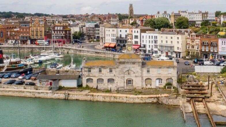 A historic harbor scene shows old buildings, cars, and boats by the water. The foreground features a stone building with a clock, while a small railway ends at the waterfront. The town rises behind with shops, homes, and a church tower.