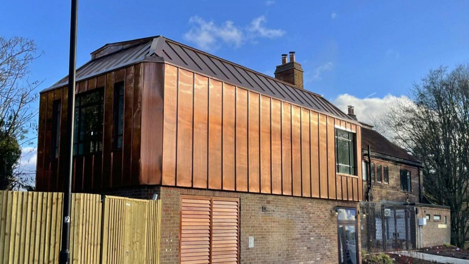 A modern house with a copper-clad upper floor and a brick lower floor, featuring large windows, a sloped roof, and set against a blue sky with scattered clouds.