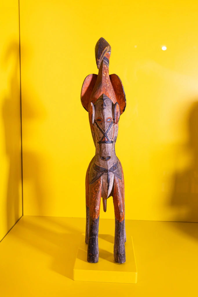 A wooden elephant sculpture with carved patterns stands on a yellow platform against a bright yellow background. The elephant has large ears and a curved trunk.