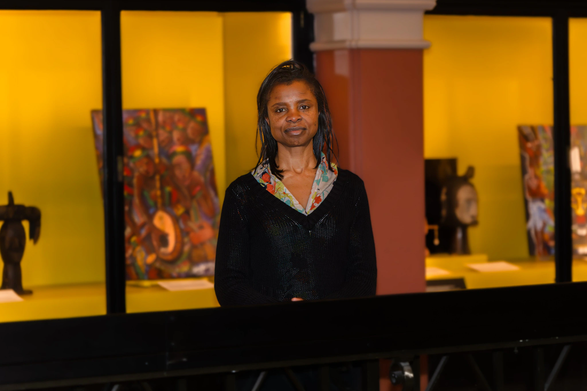 A woman with dark braided hair stands indoors in front of a yellow-lit display featuring colorful art and sculptures, looking at the camera with a gentle expression.