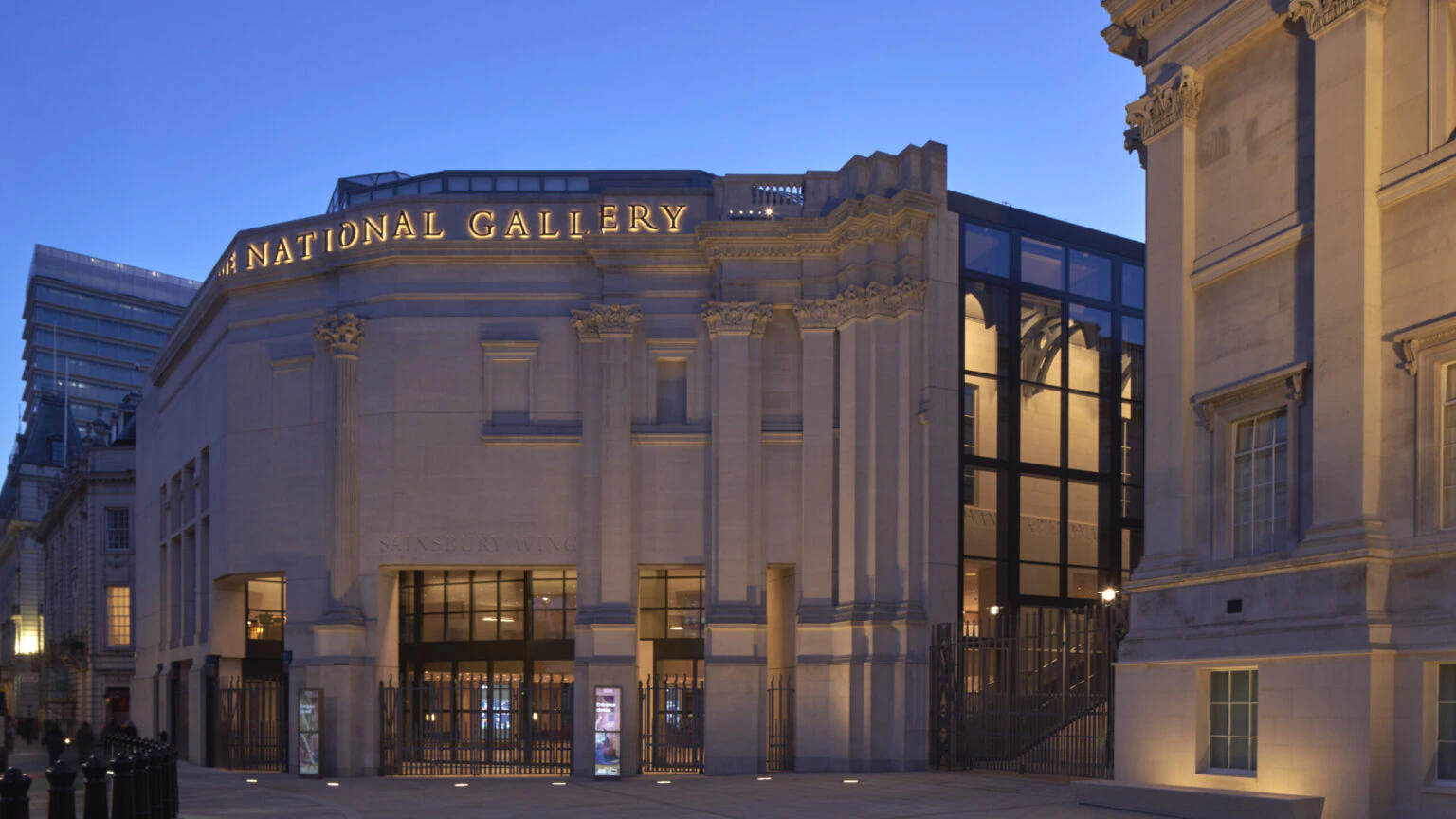 The exterior of the National Gallery at dusk, with illuminated signage and soft golden lighting highlighting the buildings classical architectural features and large glass windows.
