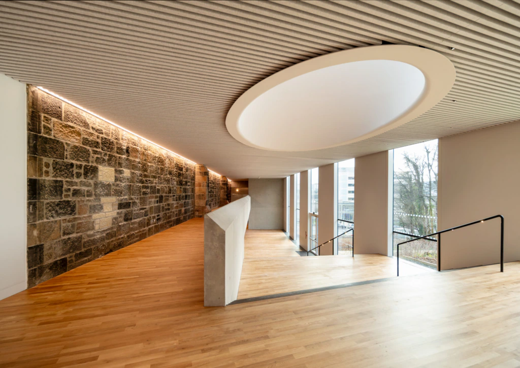 Modern interior with wooden floors, a feature stone wall on the left, large windows to the right, a sculptural concrete railing, and a ceiling with a large circular recessed light fixture.