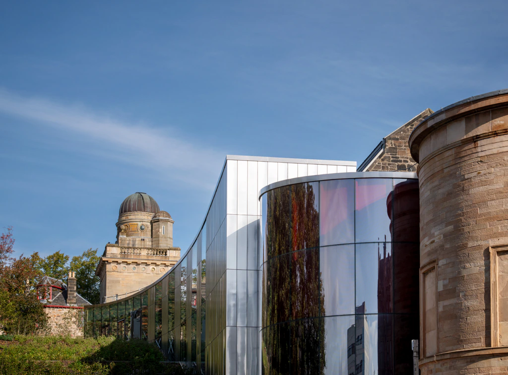 A modern glass building with curved walls stands between two historic stone buildings, including an old observatory with a domed roof, under a clear blue sky. Trees are reflected in the glass facade.