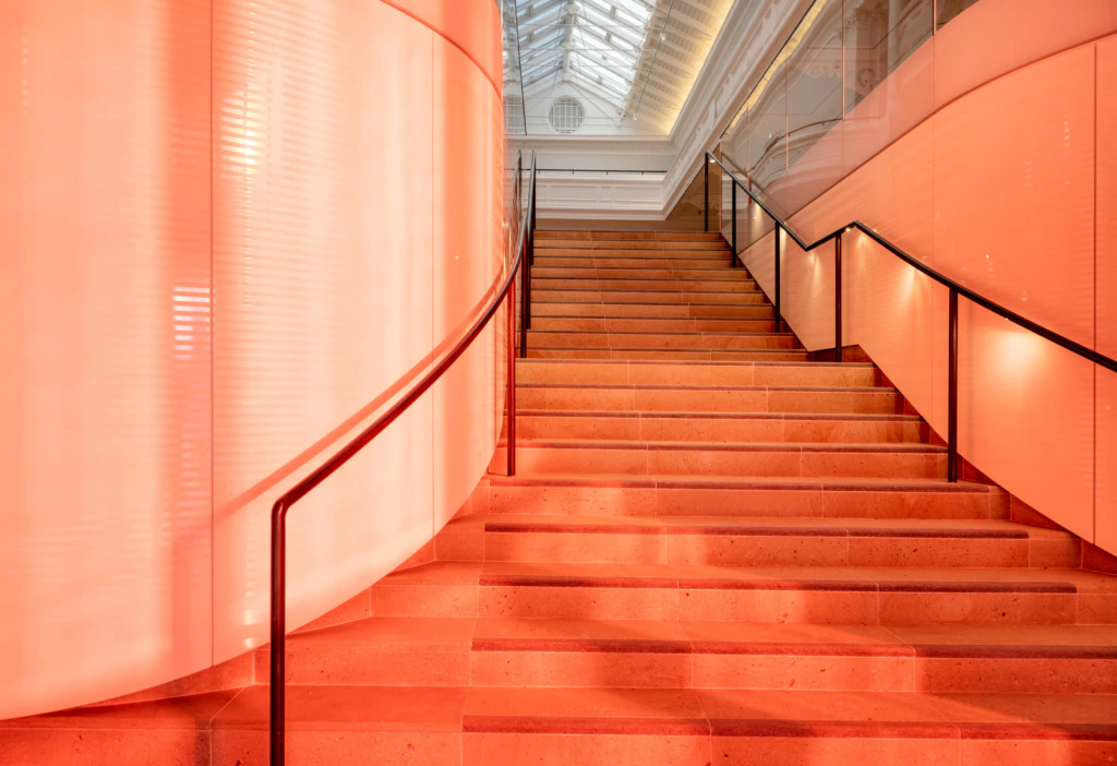 A modern staircase with orange-tinted steps and black railings ascends between curved and straight walls, with natural light streaming in from a glass ceiling above.