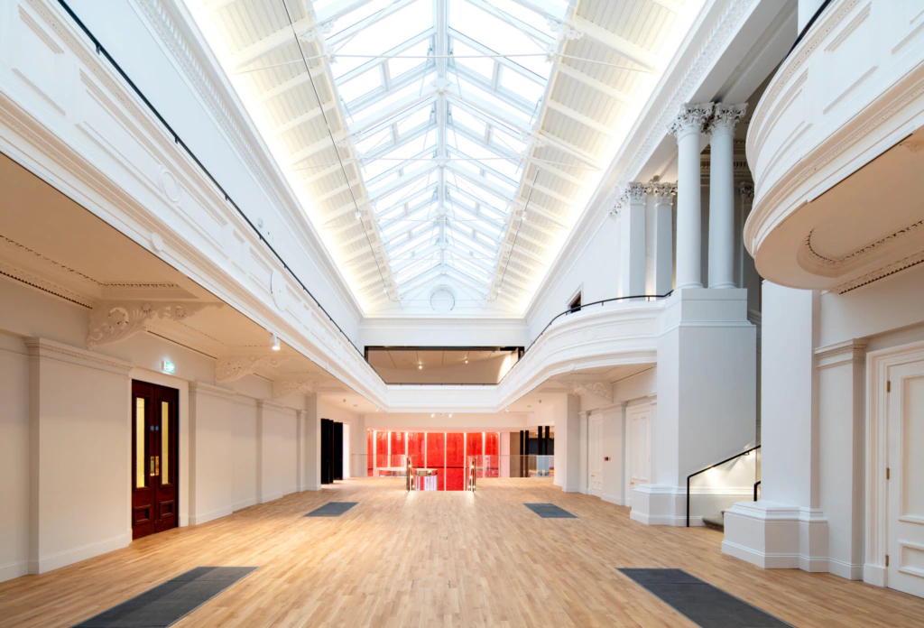 A spacious, bright hall with a high glass ceiling, white walls, classical columns, and wooden floors. Red doors and furniture are visible at the far end, giving the space a modern yet historic feel.