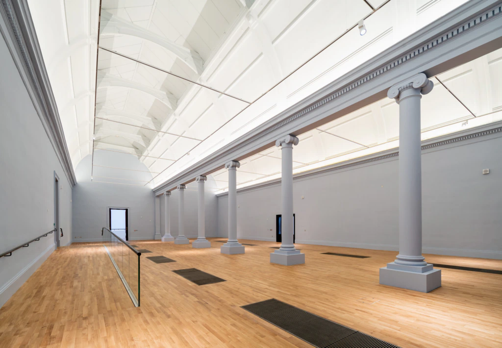 A spacious, empty gallery with light wooden floors, tall white columns, high arched ceilings, and soft natural light streaming in through skylights. Walls are plain and light-colored.