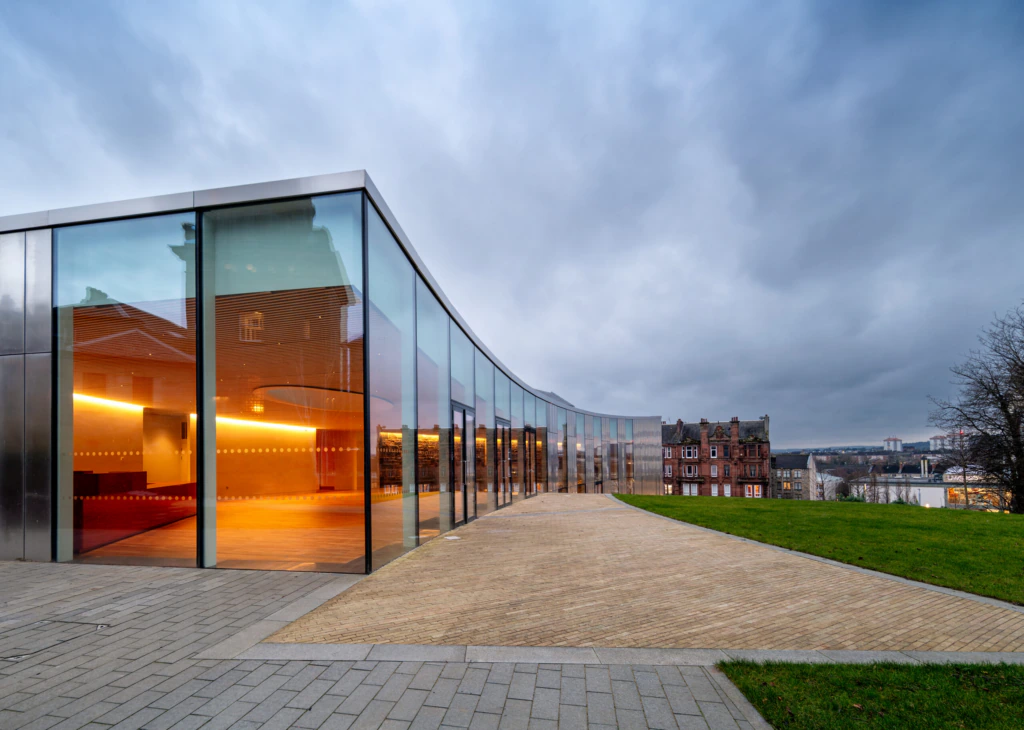 A modern glass building with curved walls reflects the cloudy sky at dusk. Warm interior lights contrast with the cool exterior. A paved path and grassy area lead to historic brick buildings in the background.