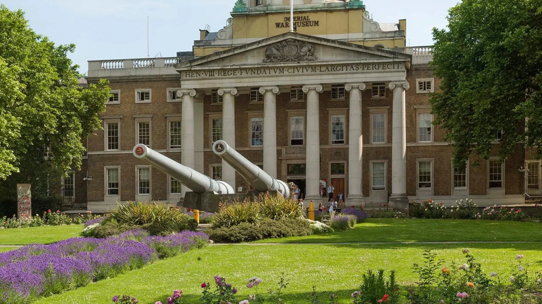 The image shows the front of the Imperial War Museum in London, with two large naval guns on the lawn, surrounded by greenery, flowers, and people entering the building. The museum’s name is visible above the entrance.