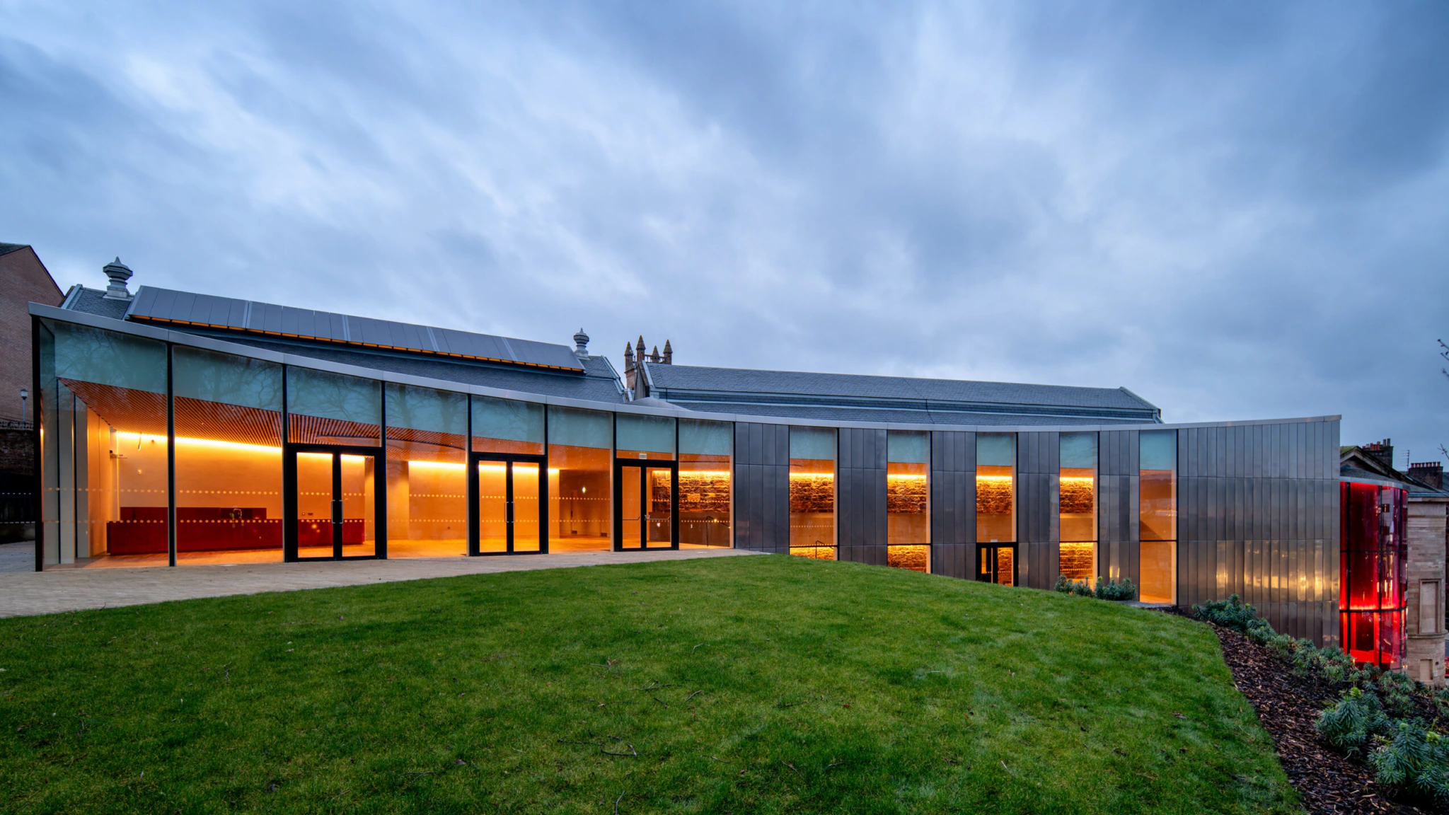 A modern building with large glass windows and metallic panels is illuminated with warm lights, contrasting against a cloudy evening sky. The structure sits on a grassy lawn with landscaped borders.
