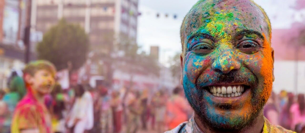 A man with a big smile stands covered in colorful powder during a vibrant outdoor festival. Other people, also covered in colors, celebrate in the blurred background.