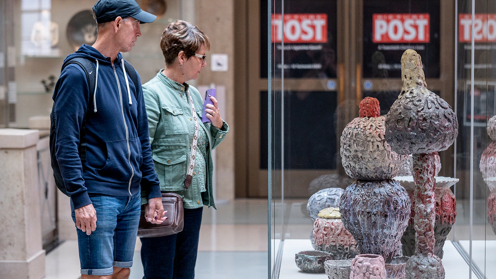 A man and a woman stand in front of a glass display case, looking at colorful, textured ceramic sculptures in a museum or gallery setting.