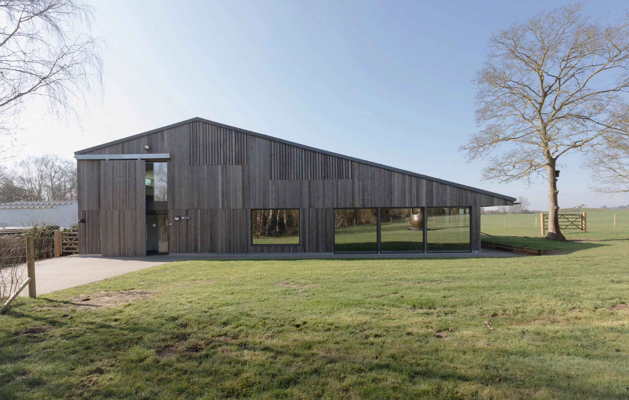 A modern building with a slanted roof and wooden exterior, featuring large glass windows, sits on a grassy lawn next to a bare tree under a clear blue sky.