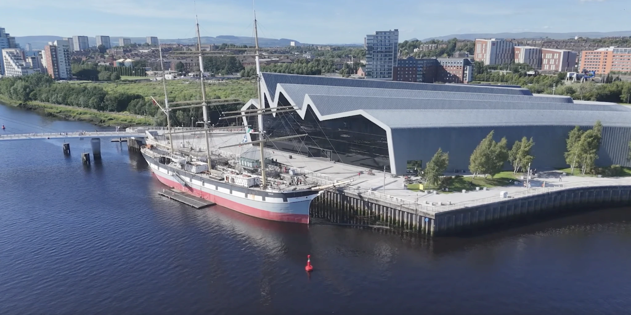 A tall ship with red trim is docked beside a modern, angular building with a zigzag roof, next to a river. City buildings, trees, and distant hills are visible in the background.