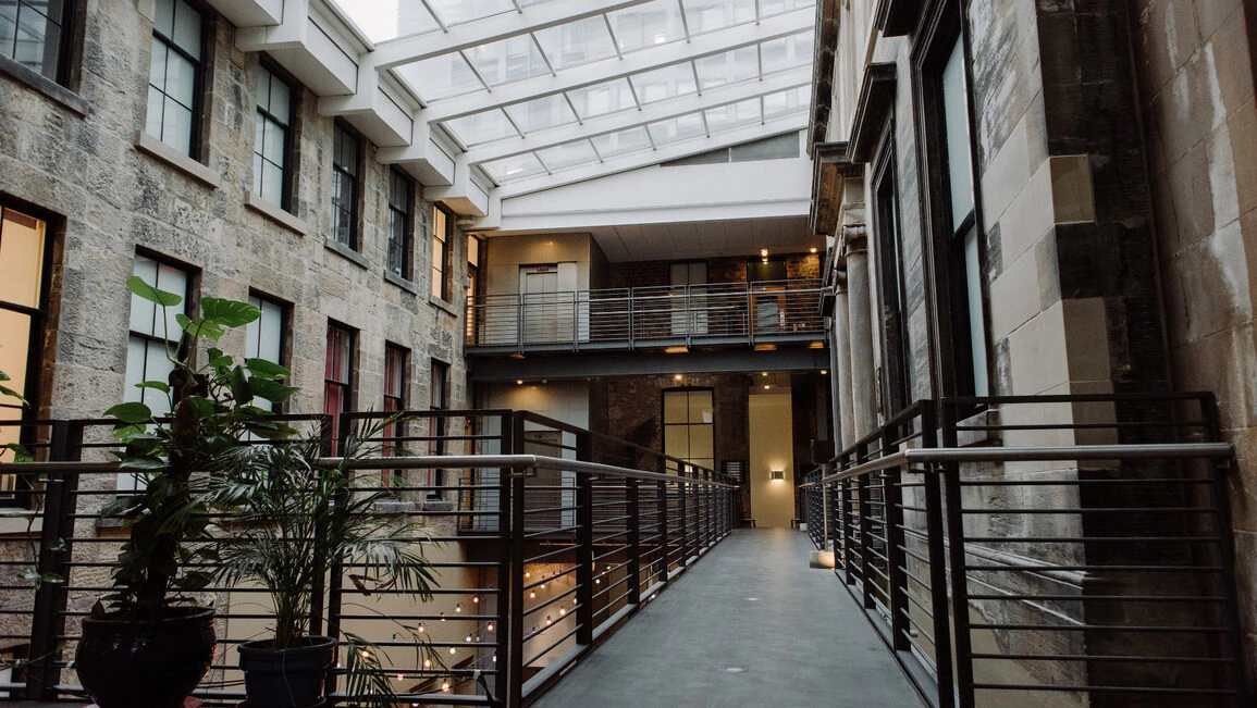 A modern indoor walkway with metal railings connects two levels of a building with stone walls, large windows, potted plants, and a glass ceiling letting in natural light.