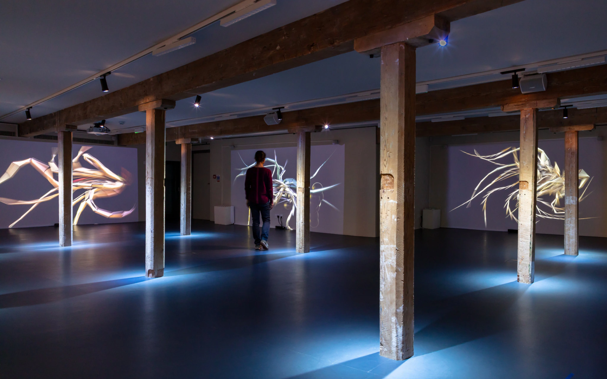 A person walks through a dimly lit gallery space with wooden pillars and digital abstract art projections displayed on the walls.