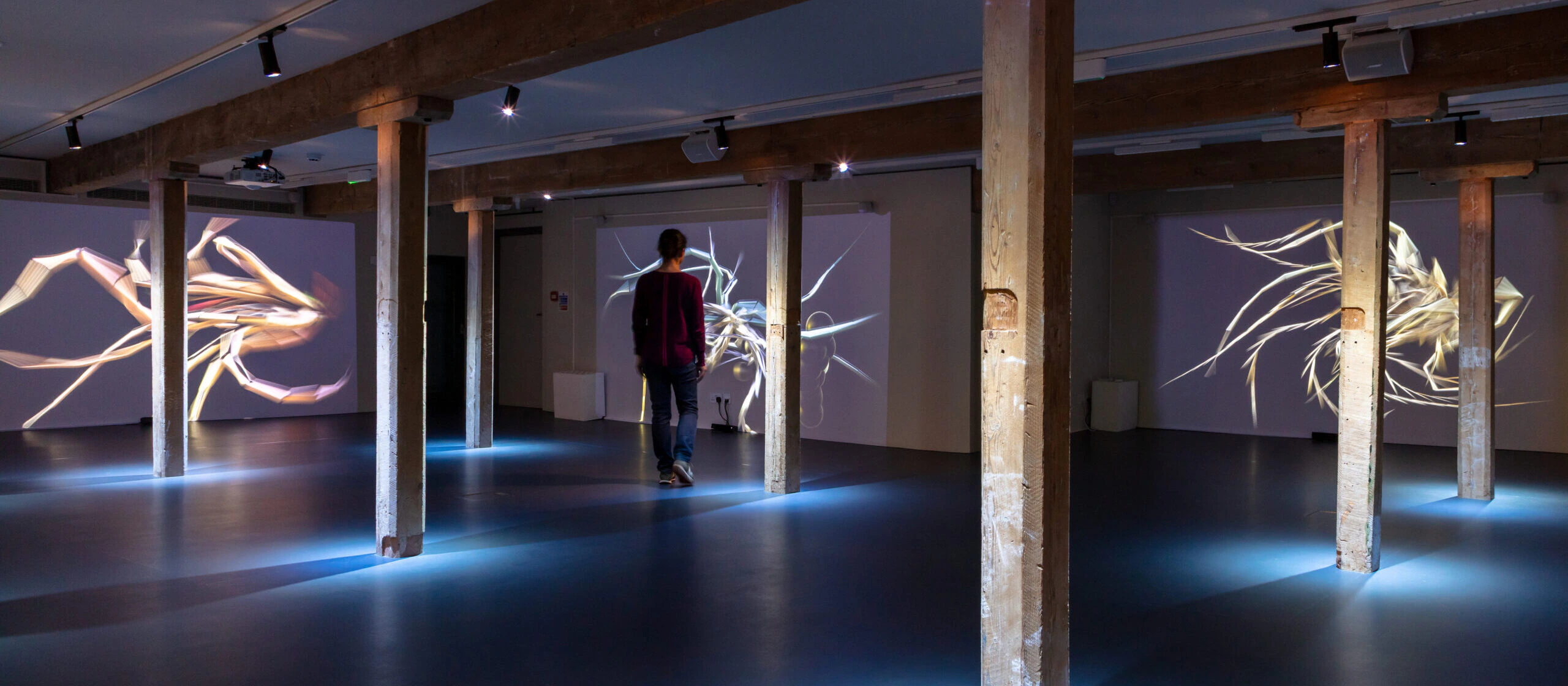 A person walks through a dimly lit gallery space with wooden pillars and digital abstract art projections displayed on the walls.