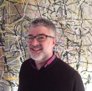 A smiling man with short gray hair, a beard, and glasses stands in front of an abstract artwork featuring intersecting colorful lines. He is wearing a dark sweater over a collared shirt.