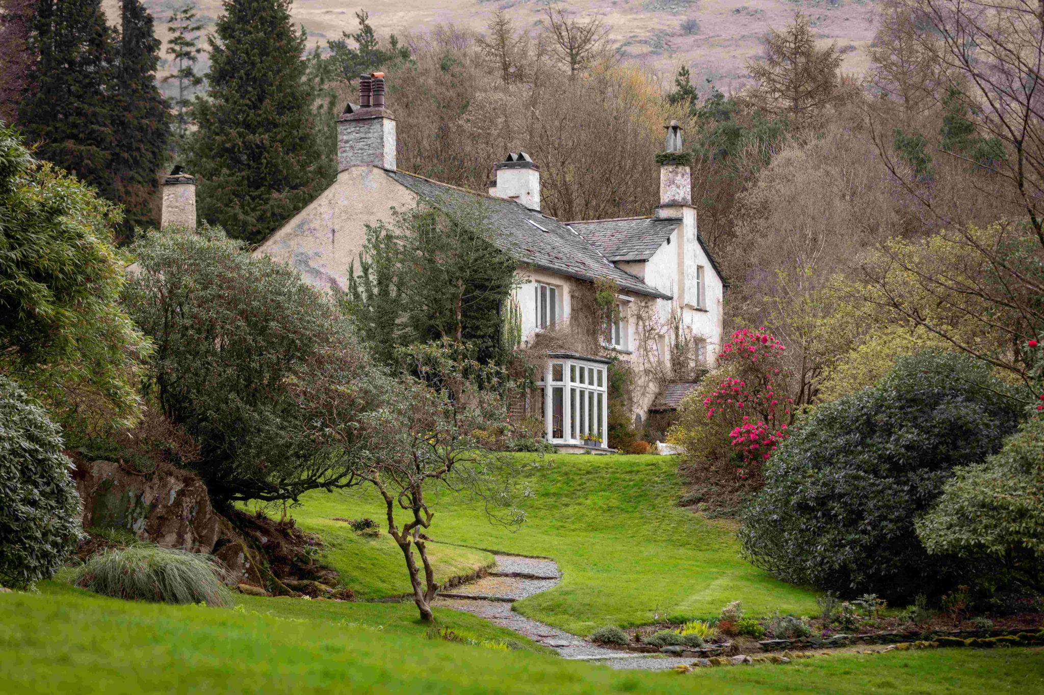 A charming, old-fashioned house with chimneys sits among lush green lawns, trees, and flowering bushes at the edge of a wooded hillside. A narrow path winds through the garden toward the house.
