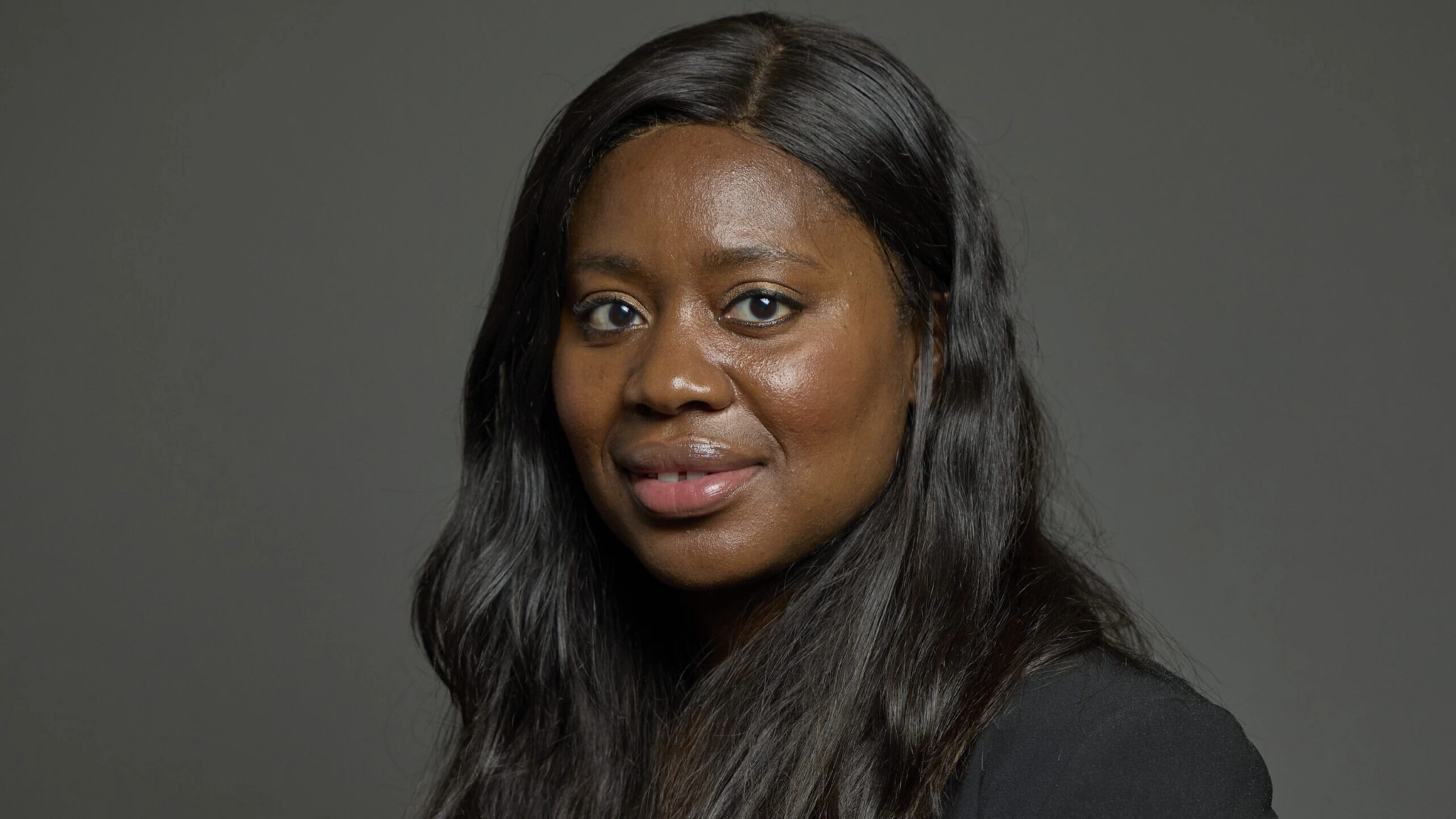A woman with long, wavy black hair wearing a black blazer poses against a plain gray background, looking directly at the camera with a slight smile.