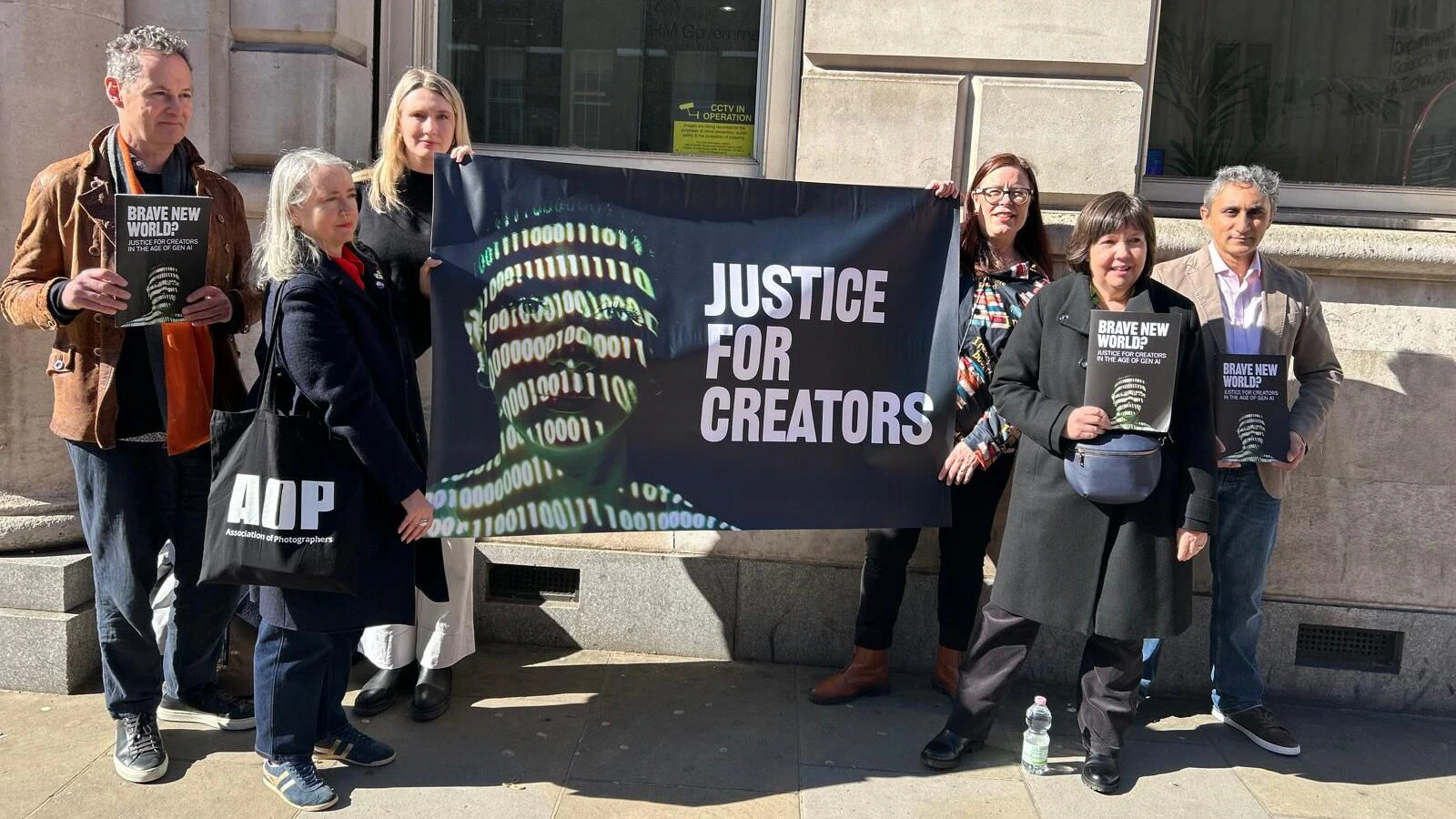 Seven people stand on a sidewalk holding a banner that reads JUSTICE FOR CREATORS with a digital face and binary code. Several hold pamphlets titled BRAVE NEW WORLD? They appear to be at an advocacy event.