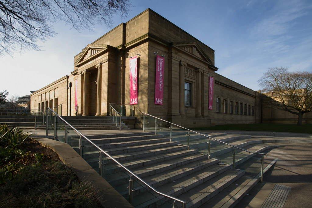 A neoclassical stone building with columns and pink banners stands behind wide steps with glass railings on a sunny day. Trees and landscaped areas surround the structure.