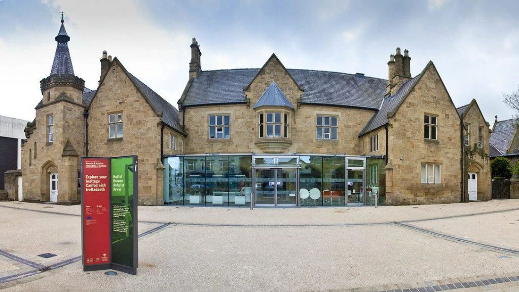 A historic stone building with gabled roofs and a turret left of center, featuring a modern glass entrance. In front, there is a tall information sign on a wide, paved area under a cloudy sky.
