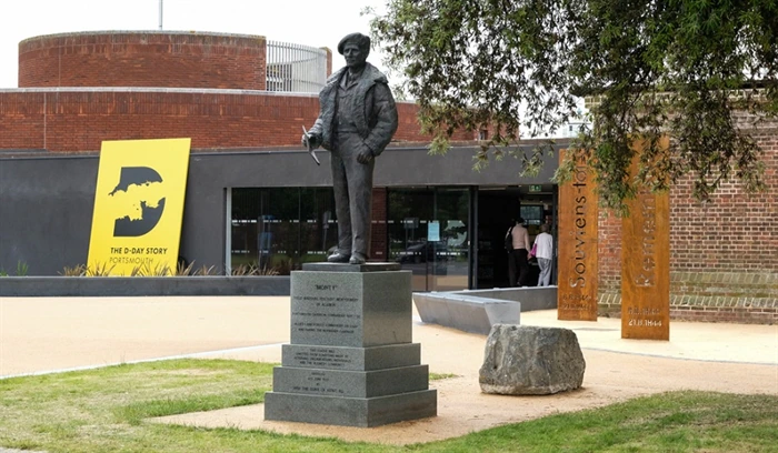 A statue stands on a stone pedestal in front of a modern building with a large yellow sign reading D-DAY STORY PORTSMOUTH. Trees and a commemorative pillar are nearby.