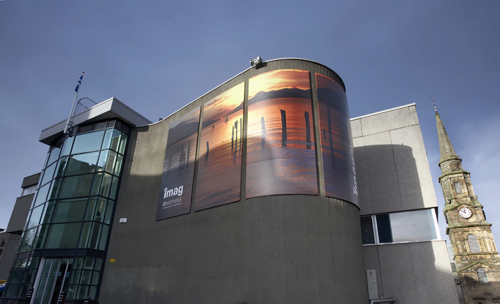 A modern building with large glass windows displays a mural of a sunset over water on its curved exterior wall, while a historic clock tower stands to the right in the background.