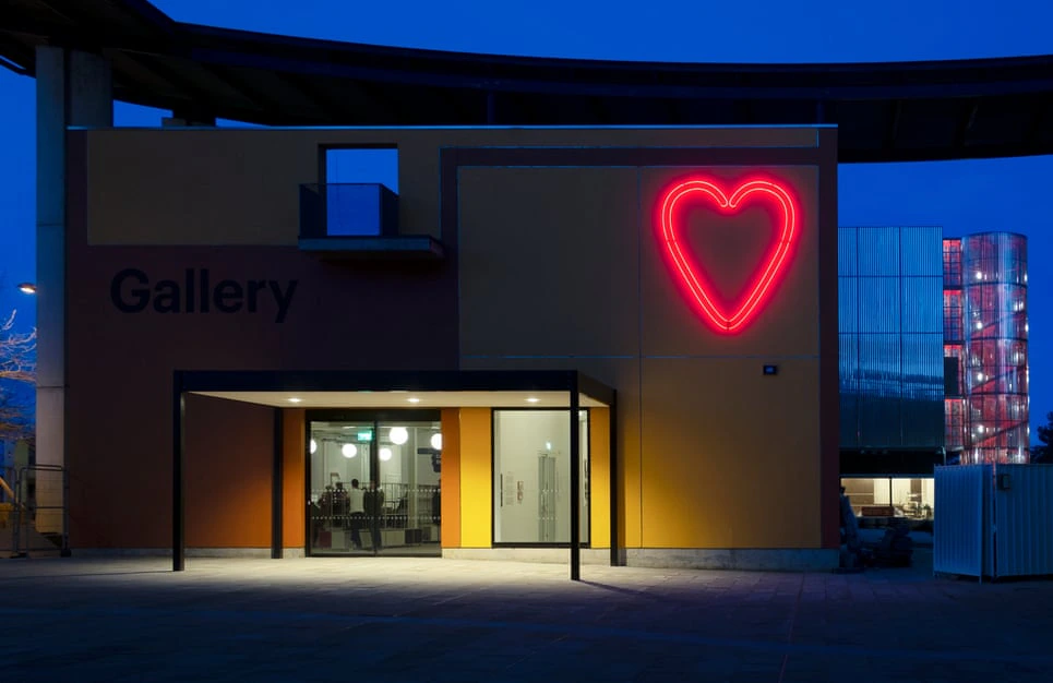 A modern gallery building at night with a large red neon heart on its wall, warm light glowing from inside, and the word Gallery displayed on the exterior.