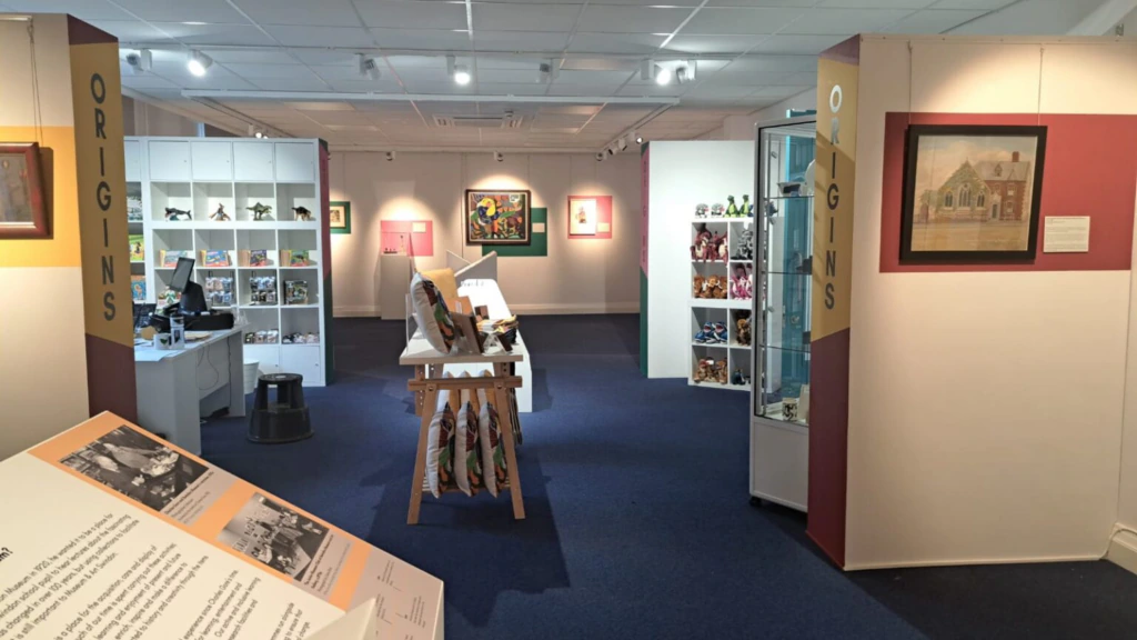 A well-lit museum exhibit room with artwork on walls, labeled “ORIGINS.” Display cases hold items, and an easel stands in the center. A person is seated at a desk in the background.