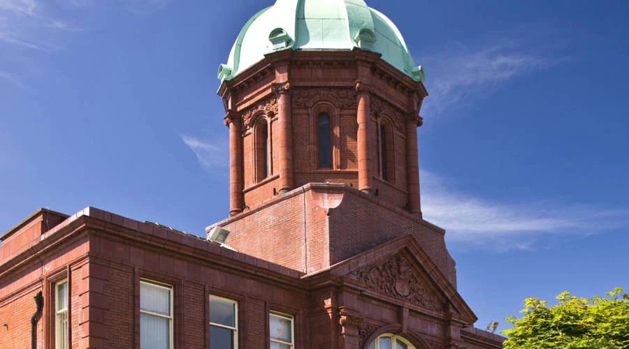 A historic red brick building with arched windows and a green copper dome tower under a clear blue sky.