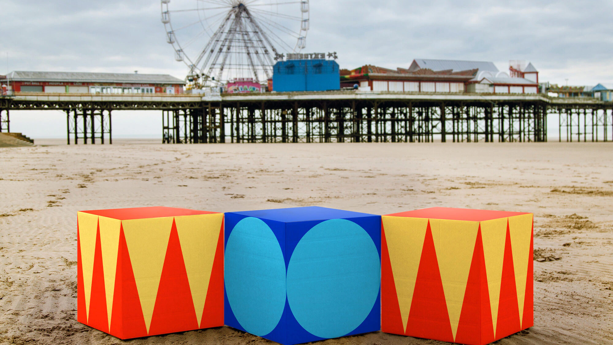 Three colorful geometric blocks sit on a sandy beach in front of a pier with a Ferris wheel and buildings in the background under a cloudy sky.
