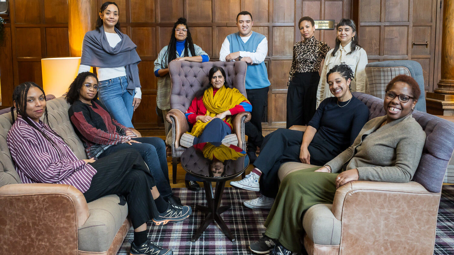 A group of ten people, diverse in gender and appearance, pose together in a wood-paneled room. Some are seated on armchairs while others stand behind them, all smiling and looking at the camera.