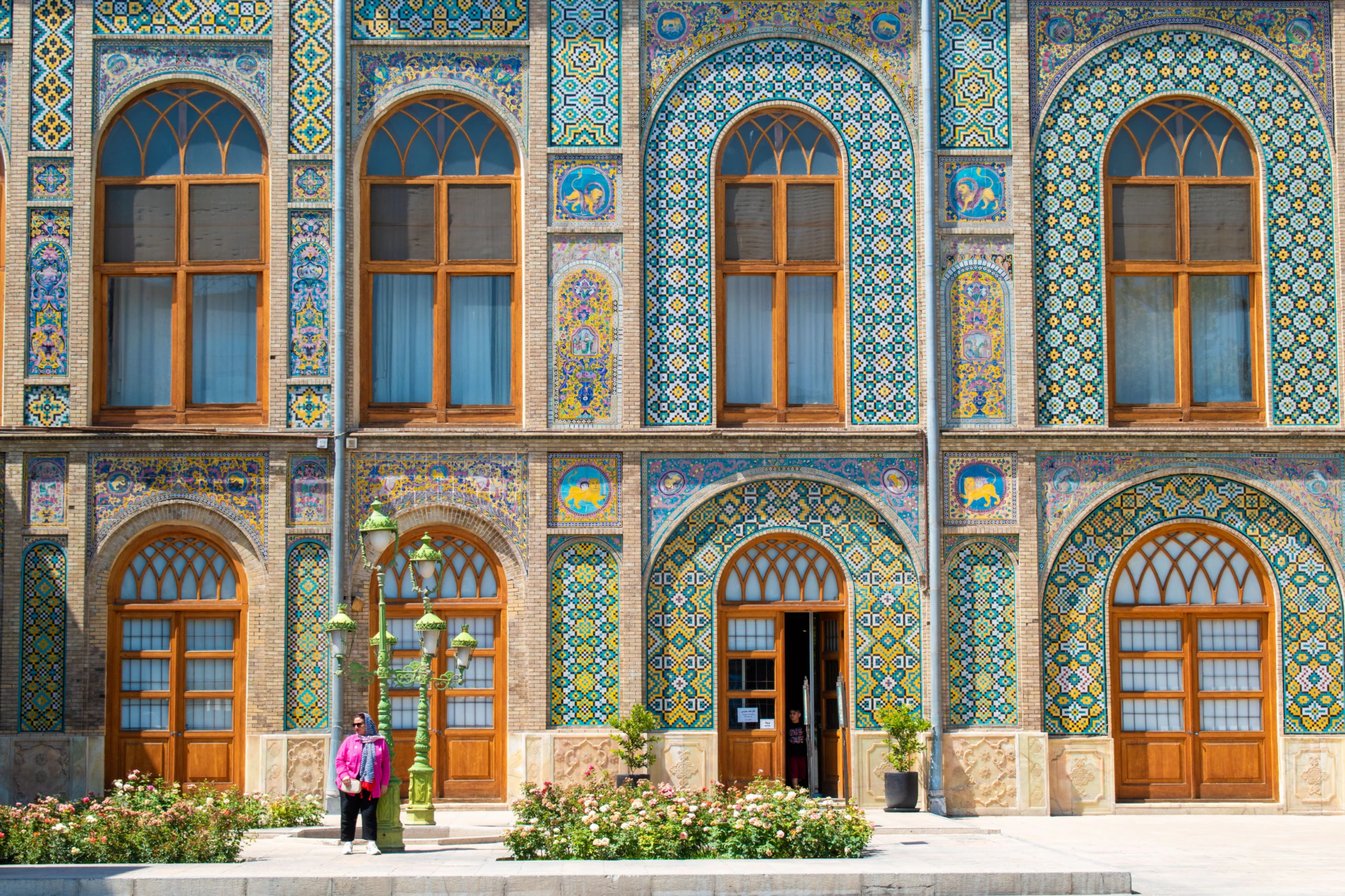 A person in a pink jacket stands in front of an ornate building facade featuring arched wooden windows and colorful, intricate tile patterns with geometric and floral designs.