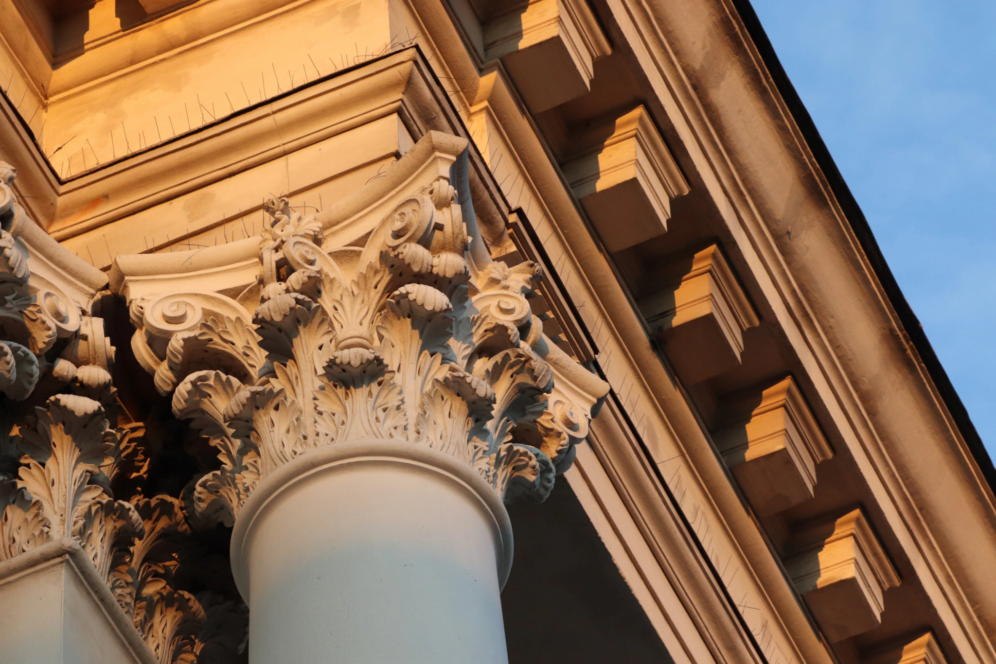 Close-up of ornate Corinthian columns and detailed architectural carvings on a building, illuminated by warm sunlight, with a blue sky in the background.