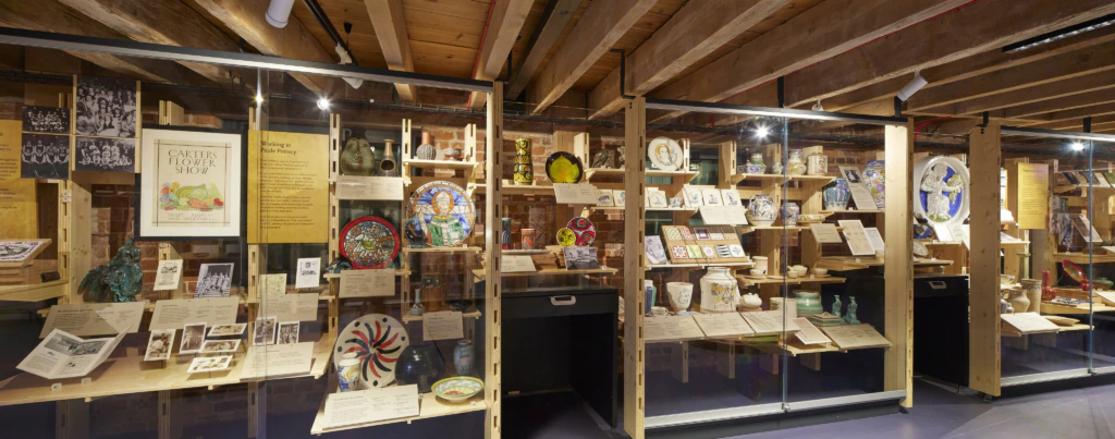 A museum exhibit displays colorful ceramic plates, pottery, historical photos, posters, and documents in glass cases on wooden shelves beneath exposed wooden beams.