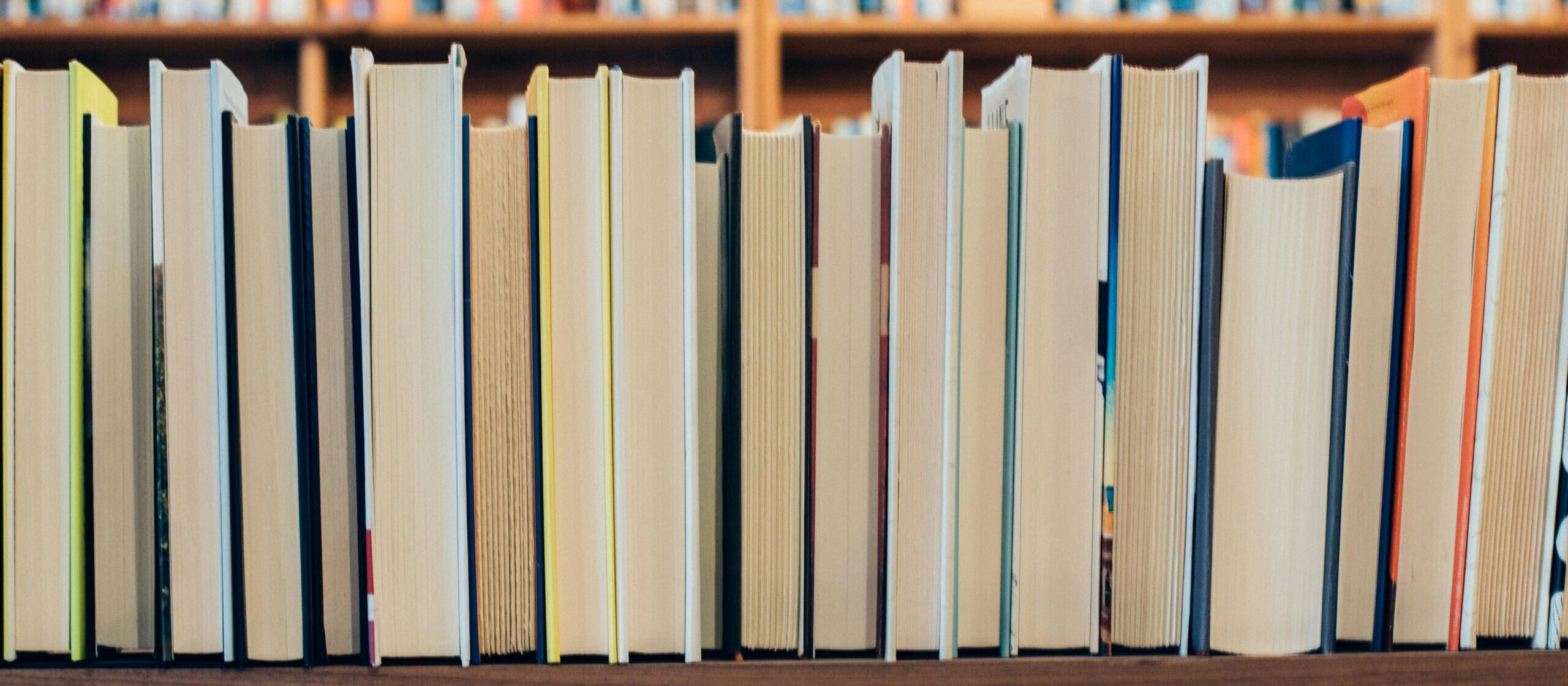 A row of upright books is seen from the side, displaying their pages rather than covers, with shelves full of more books blurred in the background.