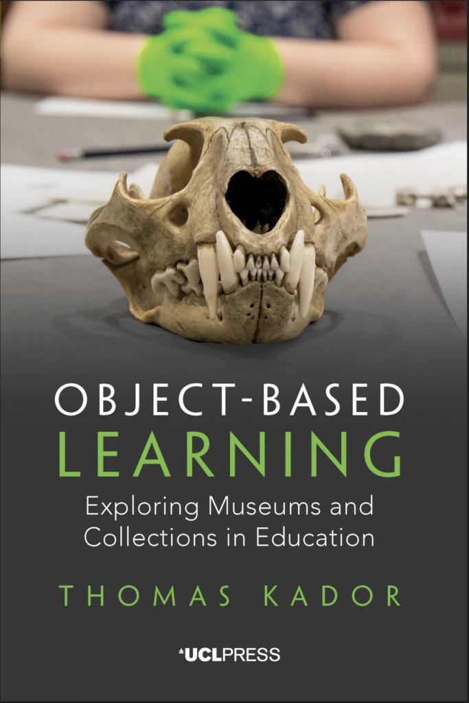 Book cover showing an animal skull with large teeth on a table, paper and writing tools nearby, and a person in the background wearing green gloves. Title: “Object-Based Learning” by Thomas Kador.