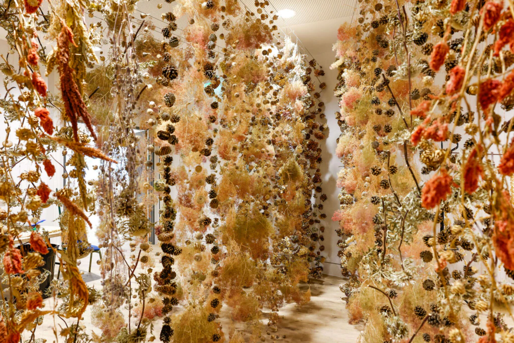 Hanging installations made of dried flowers, plants, and pine cones fill a bright indoor space, creating a natural, forest-like atmosphere along a wooden floor.