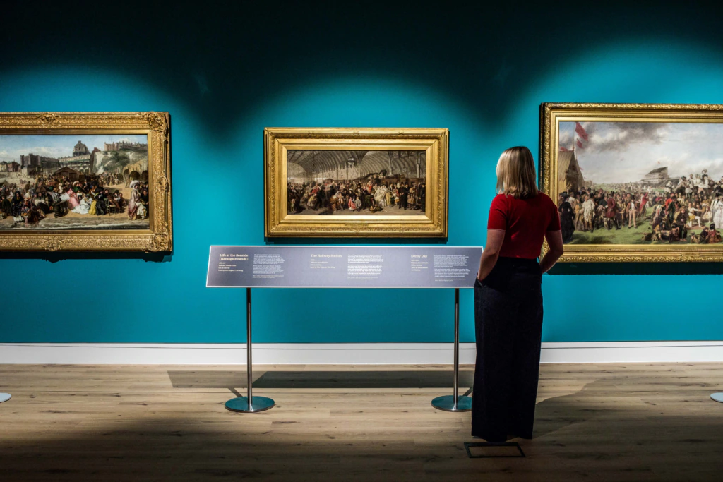 A woman stands in an art gallery, viewing three large framed historical paintings on a teal-colored wall, with an information panel in front of her. The gallery has wooden floors and bright lighting.
