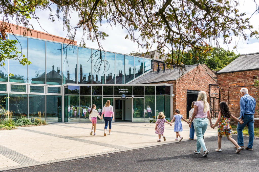 A group of adults and children walk hand-in-hand toward a modern glass building with Museum Entrance on the door, surrounded by trees and brick structures on a sunny day.