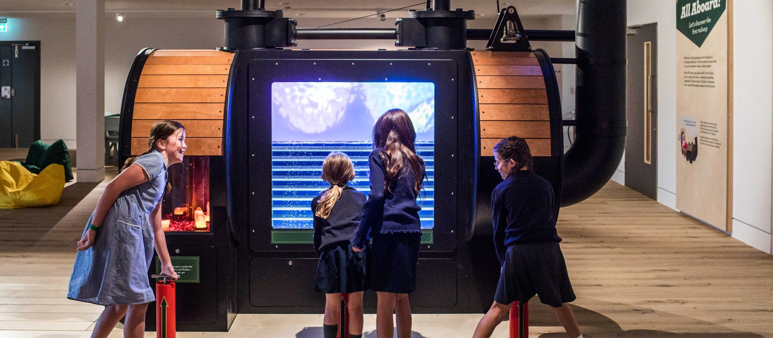Four children interact with a large digital display shaped like a wooden barrel in a museum exhibit. They appear engaged and curious, using red levers attached to the display. The room is brightly lit with wooden floors.