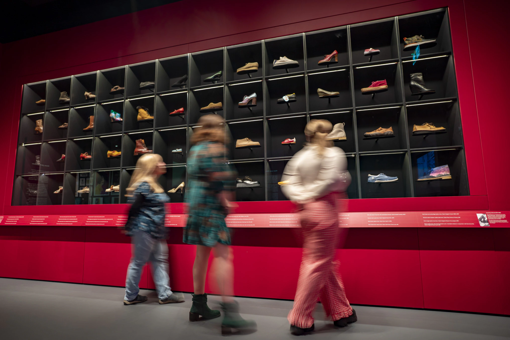Three people walk past a red wall display featuring dozens of shoes in individual cubbies, showcasing a variety of styles and colors in a museum or exhibit setting.