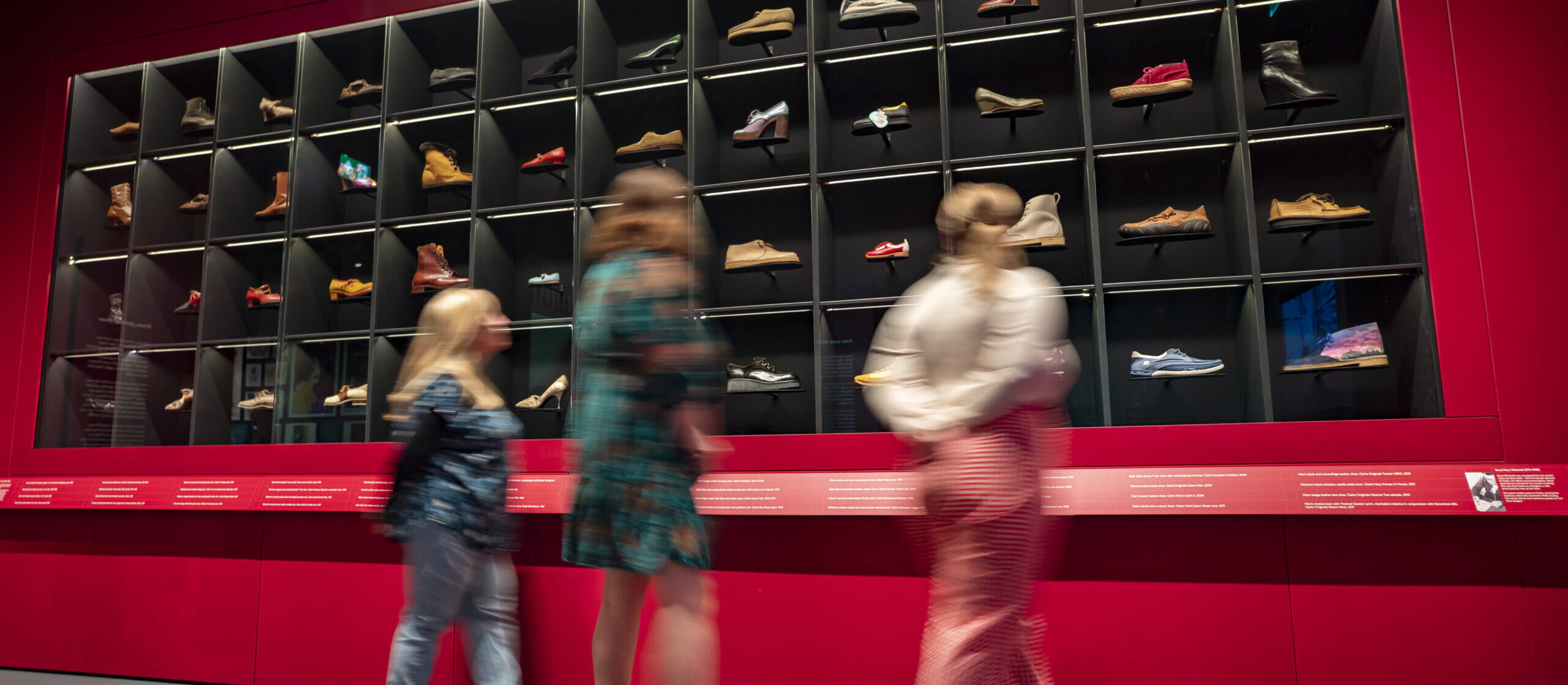 Three people walk past a red wall display featuring dozens of shoes in individual cubbies, showcasing a variety of styles and colors in a museum or exhibit setting.