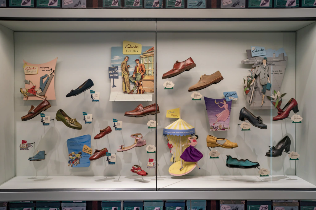 A display case showcases various vintage Clarks shoes with colorful, retro advertisements and labels, arranged creatively with some shoes elevated or paired with illustrated cutouts and 3D decorative elements.