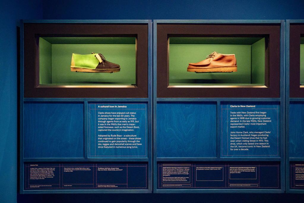 Two pairs of shoes are displayed in glass cases against a green background with blue walls. Informational plaques beneath each case describe the significance of the shoes.