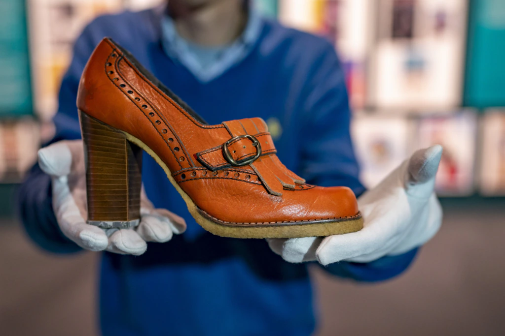A person wearing white gloves holds up an orange vintage high-heeled shoe with a buckle and decorative stitching, with a blurred background of colorful shelves or posters.