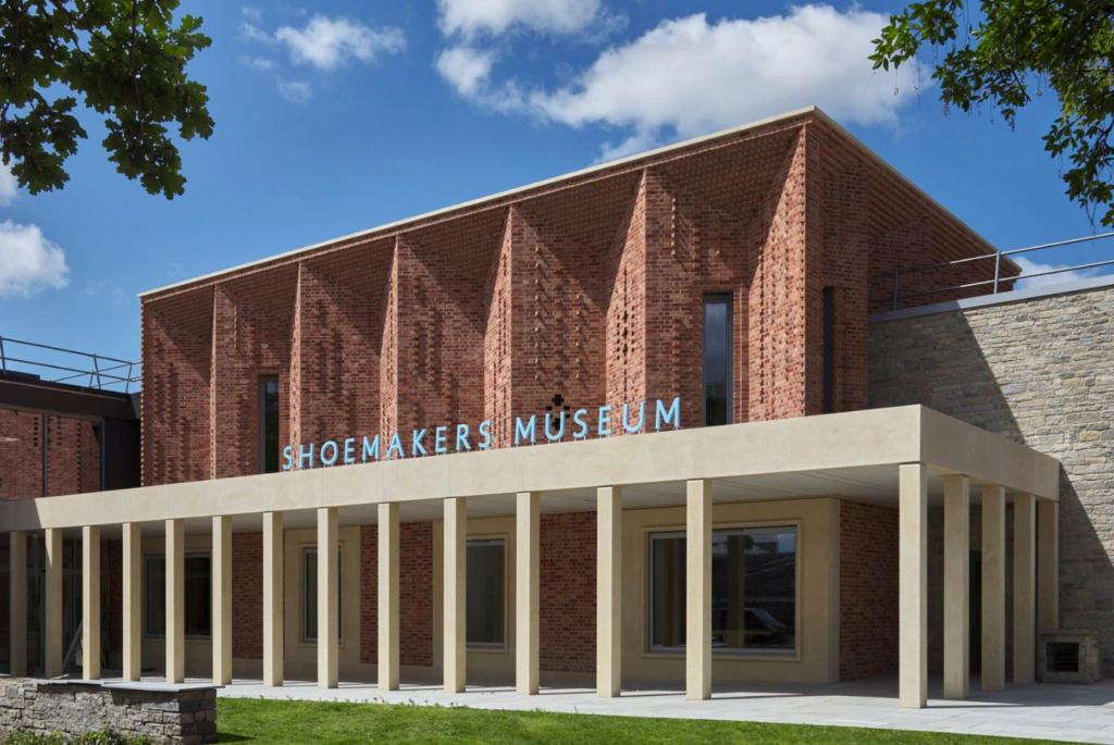 A modern brick building with vertical brick columns, large windows, and a beige portico. Blue letters on the facade read Shoemakers Museum. The sky is clear with some clouds, and trees partially frame the image.