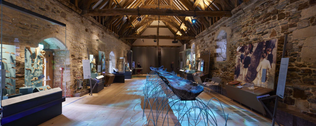 A museum gallery with stone walls and a high, wooden-beamed ceiling displays historical artifacts, glass cases, and a large wooden boat centerpiece under blue lighting. Exhibits with text and images line both walls.